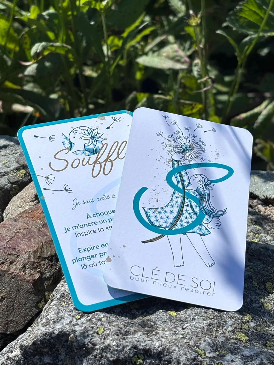 Deux cartes Souffle du jeu Clé de Soi, au liseré turquoise, posées sur un rocher : l'illustration d'un pissenlit soufflé et l'exercice au dos